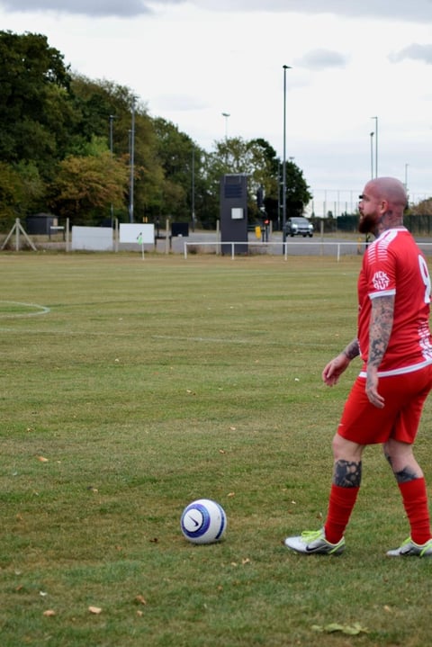 Footballer in red jersey standing on grass field next to soccer ball with stadium in background