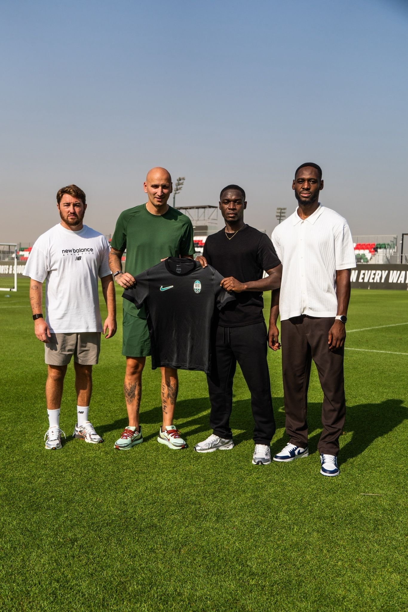 Four men standing on a soccer field holding a black jersey, with stadium and clear sky in the background