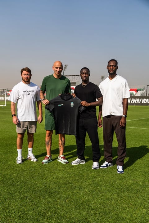 Four men standing on a soccer field holding a black jersey, with stadium and clear sky in the background